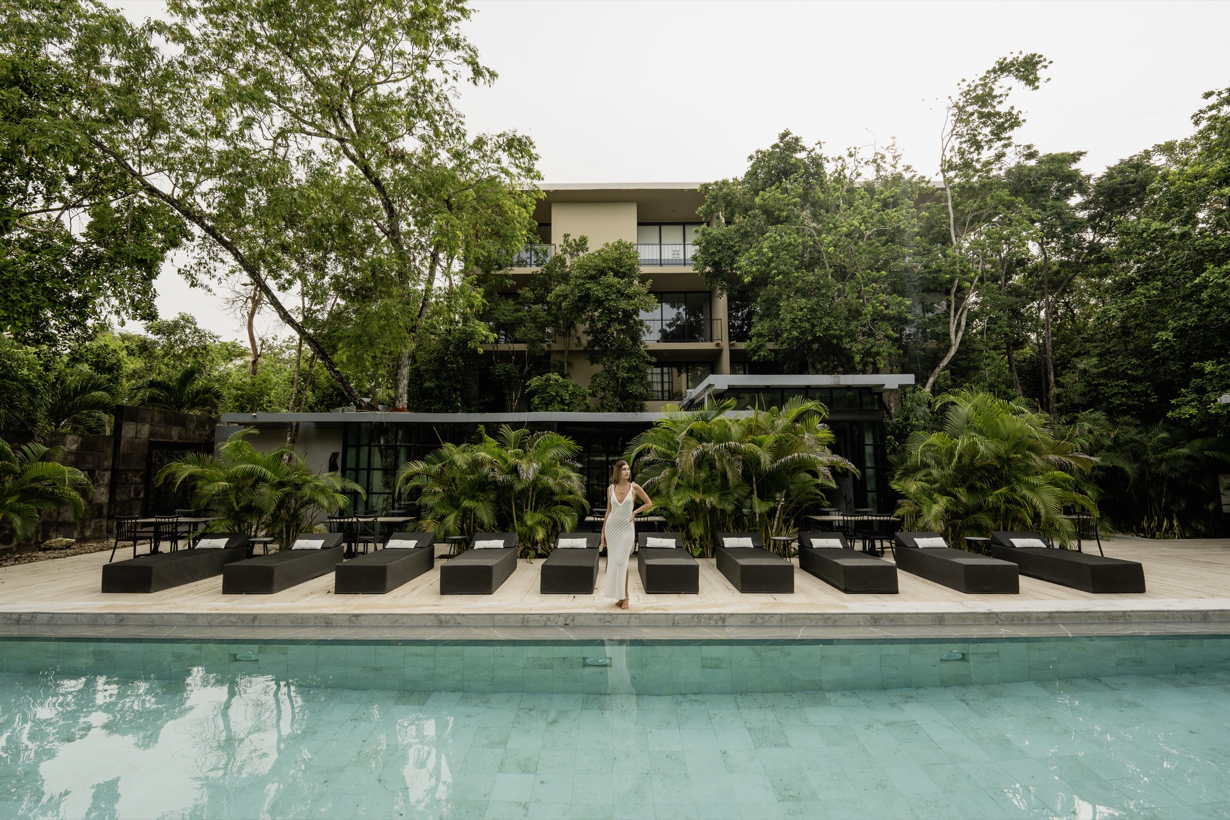Photograph of a pool with rows of black loungers, palms and a four-level building behind, a woman in a white dress standing among the loungers
