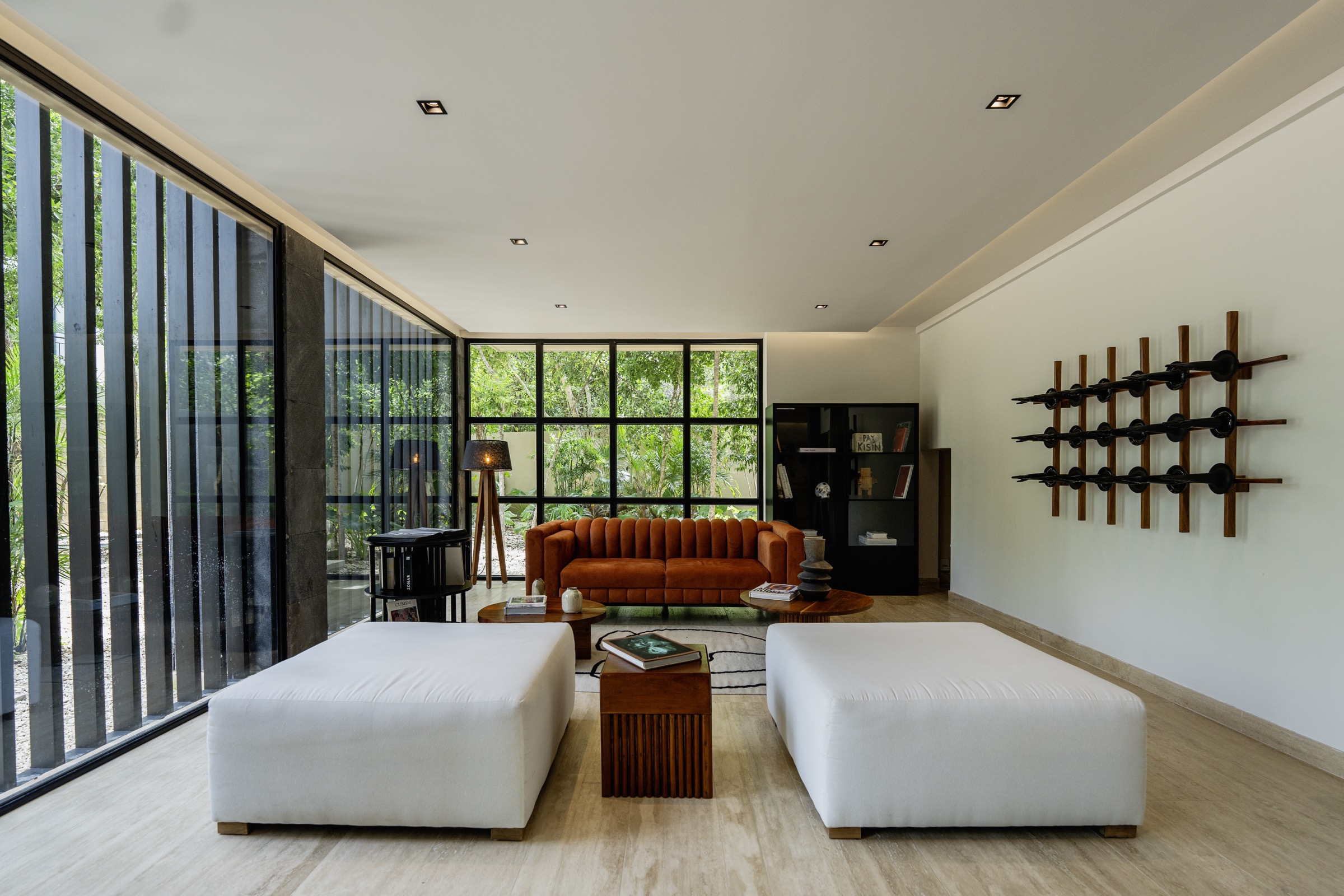 Photograph of a lobby with travertine floor, floor-to-ceiling black glazing onto a garden, a rust velvet sofa, two white ottomans and a wall piece of wooden bars with black ends