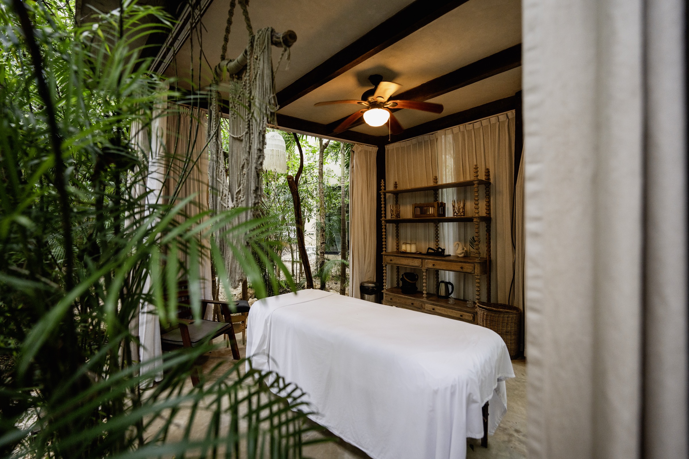 Photograph of an open-air massage cabana under a ceiling fan, with a bobbin-turned wooden shelf, linen curtains and jungle at its edge