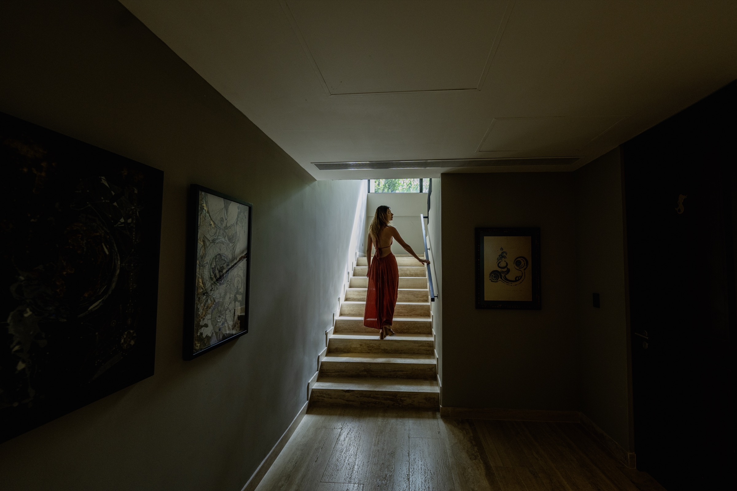 Photograph of a dark corridor with framed works on the walls and a stone stair rising toward daylight, a woman in a rust dress climbing it