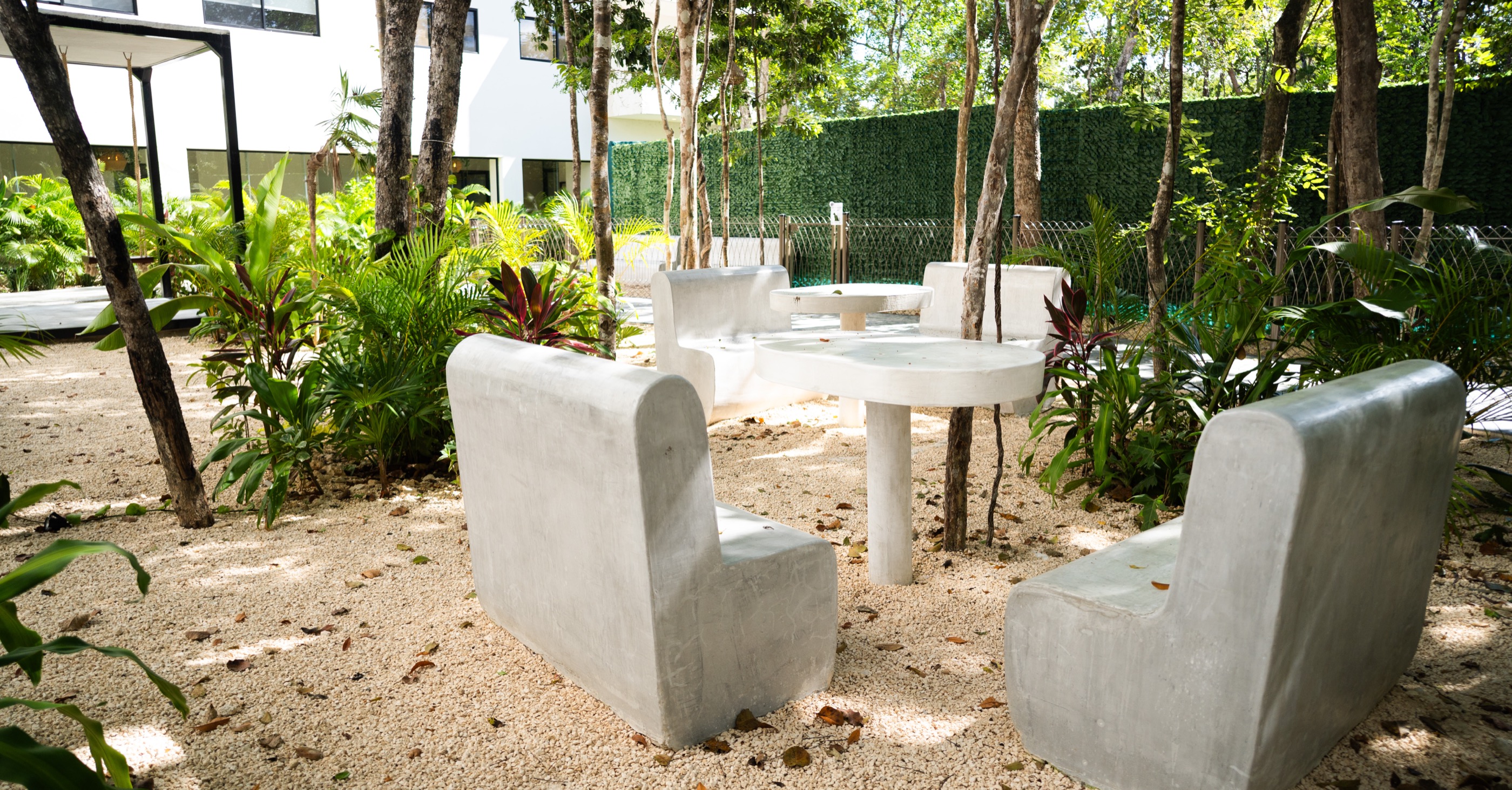 Cast-stone seating and tables on sand between the buildings, under preserved trees